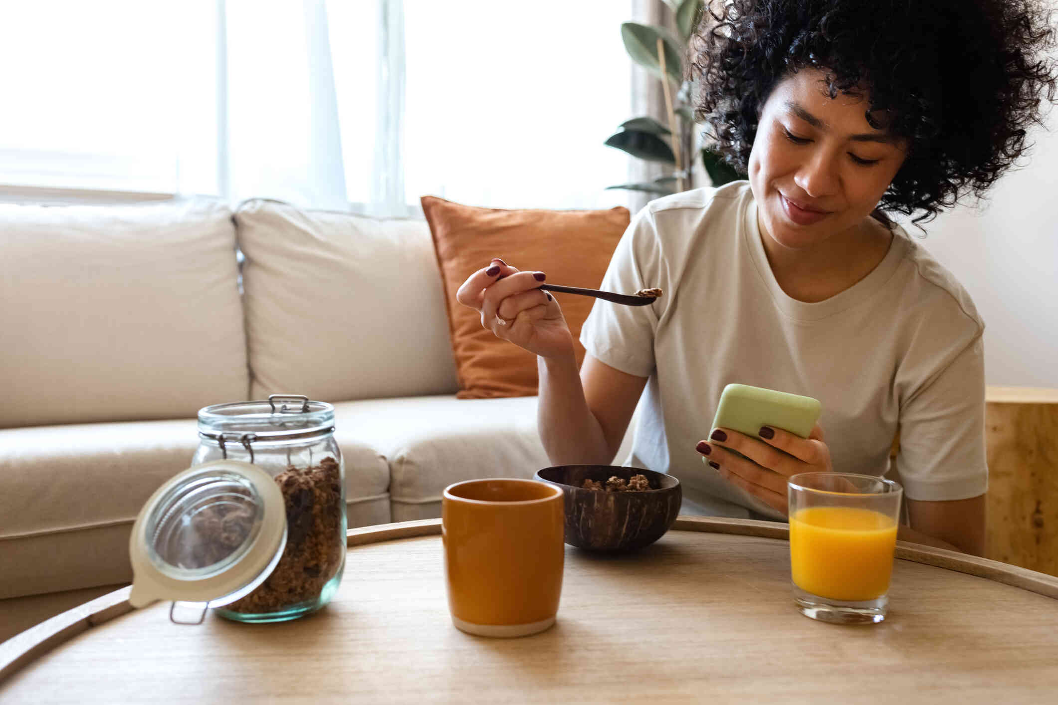 A woman sits at the kitchen table and eats from a bowl with a spoon while smiling down at the cellphone in her hand.